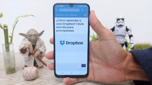 Una mano sosteniendo un teléfono inteligente que muestra una página web titulada "¿Cómo aprender a usar Dropbox? | Guía sencilla para principiantes," con el logotipo de Dropbox