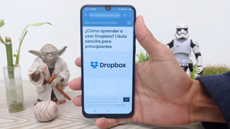 Una mano sosteniendo un teléfono inteligente que muestra una página web titulada "¿Cómo aprender a usar Dropbox? | Guía sencilla para principiantes," con el logotipo de Dropbox