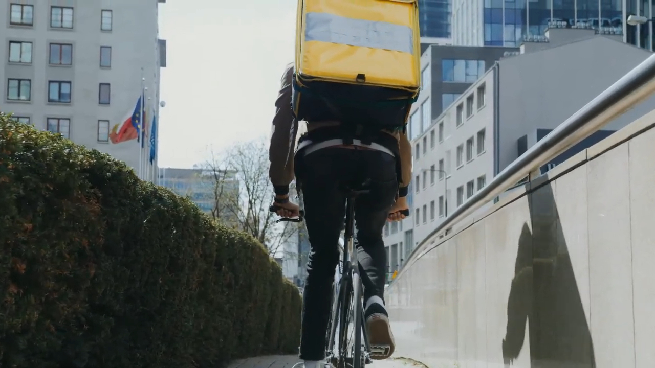 Hombre montando una bicicleta con una mochila amarilla de Rappi, pedaleando por una calle rodeada de edificios modernos y banderas