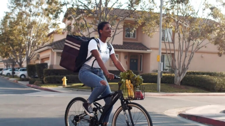 Mujer montando una bicicleta con una mochila de entrega y un cesto lleno de frutas y verduras, pasando por un vecindario residencial