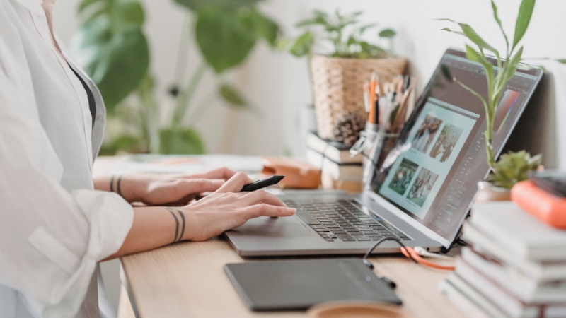 A person working on a laptop at a desk with plants, books, and design tools nearby