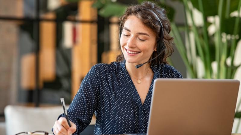 A smiling customer service representative wearing a headset takes notes while working on a laptop