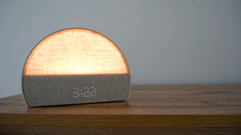 A modern sunrise alarm clock with a soft glowing light sits on a wooden nightstand