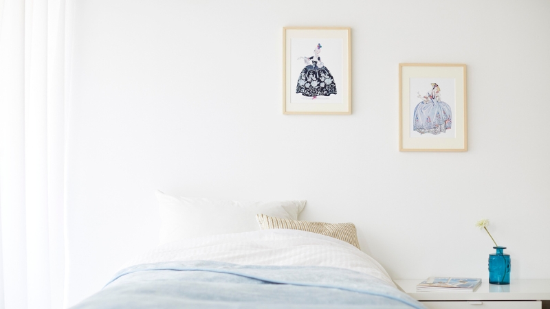 A minimal bedroom with framed art above the bed, white bedding, and a blue vase on the nightstand
