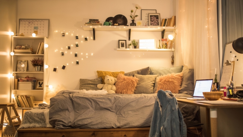 A cozy bedroom with string lights, shelves, and a bed decorated with pillows and a teddy bear