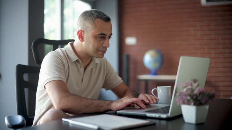 A man works on a laptop at a desk with a notebook, coffee cup, and plant nearby