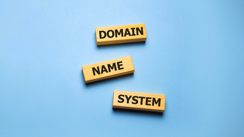 Bloques de madera con las palabras “Domain”, “Name” y “System” sobre fondo azul, representando el sistema de resolución DNS
