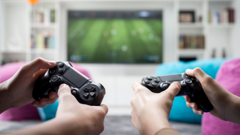 Two people hold game controllers while a football match plays on a TV