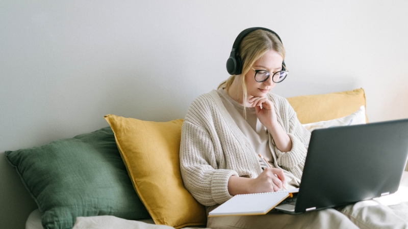 A woman with headphones studies on a laptop and writes notes while seated on a bed