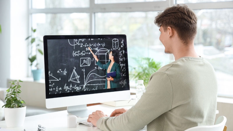 A student watches an online lesson on a computer with a teacher explaining problems in science and math on a chalkboard