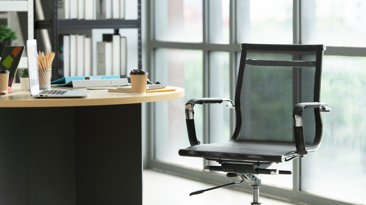 A flexible office desk area with a black chair beside large windows